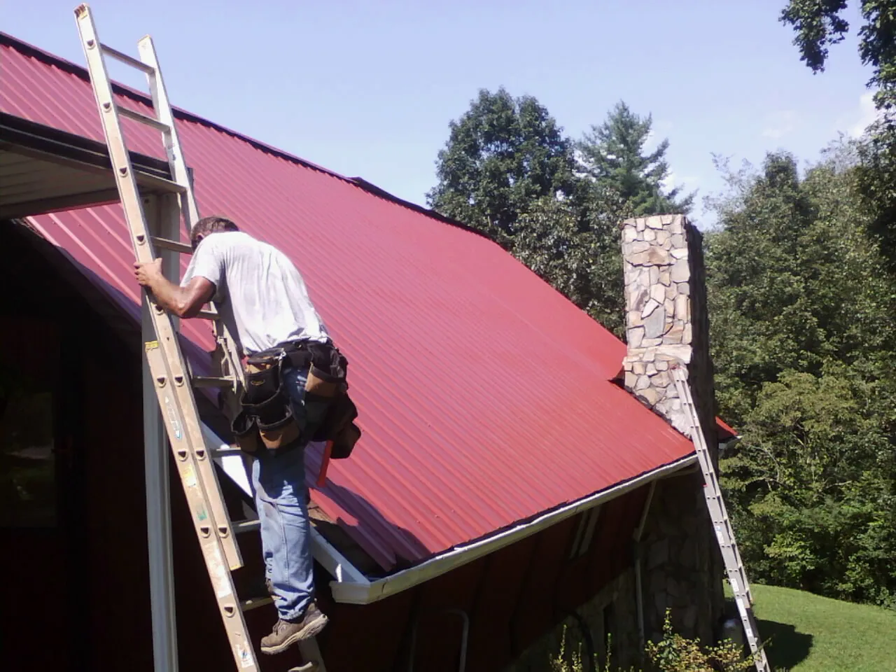 Roofer on a ladder beside a red metal roof and stone chimney — Soffit & Fascia Repair by Roofing Brothers in West Columbia